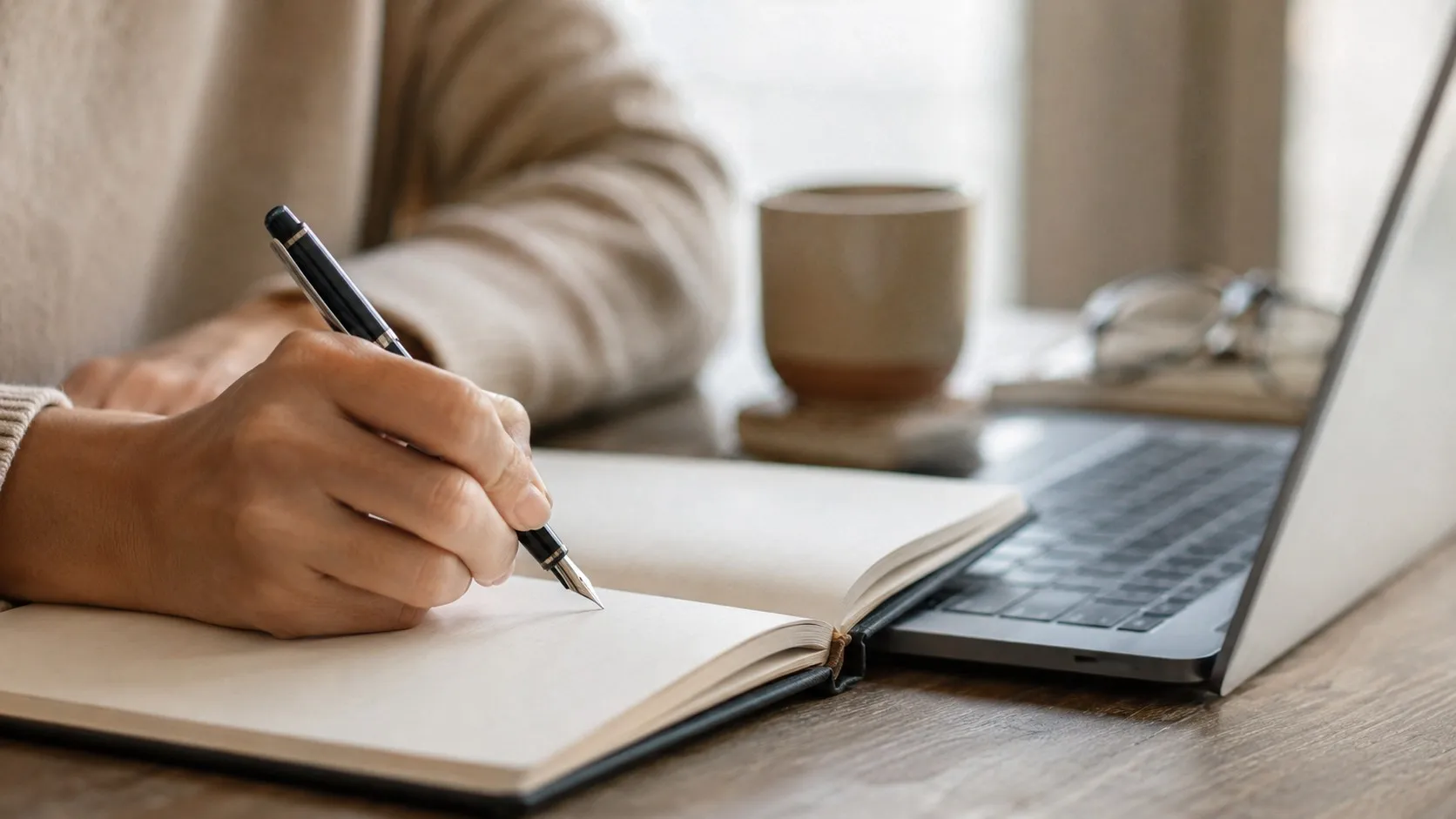 En hand skriver med bläckpenna i ett uppslaget anteckningsblock bredvid en öppen laptop och en kaffekopp