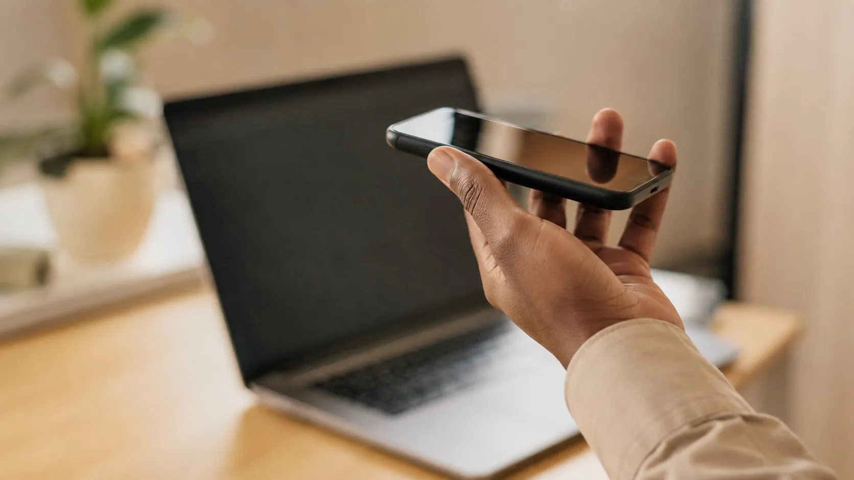 En hand håller upp en smartphone som om personen pratar in i den, en laptop oskarp i bakgrunden