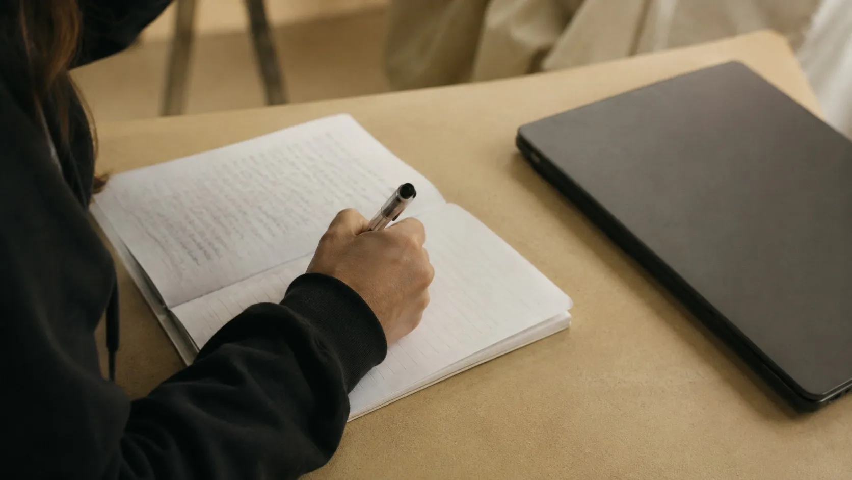 En elev skriver för hand i ett anteckningsblock bredvid en stängd laptop