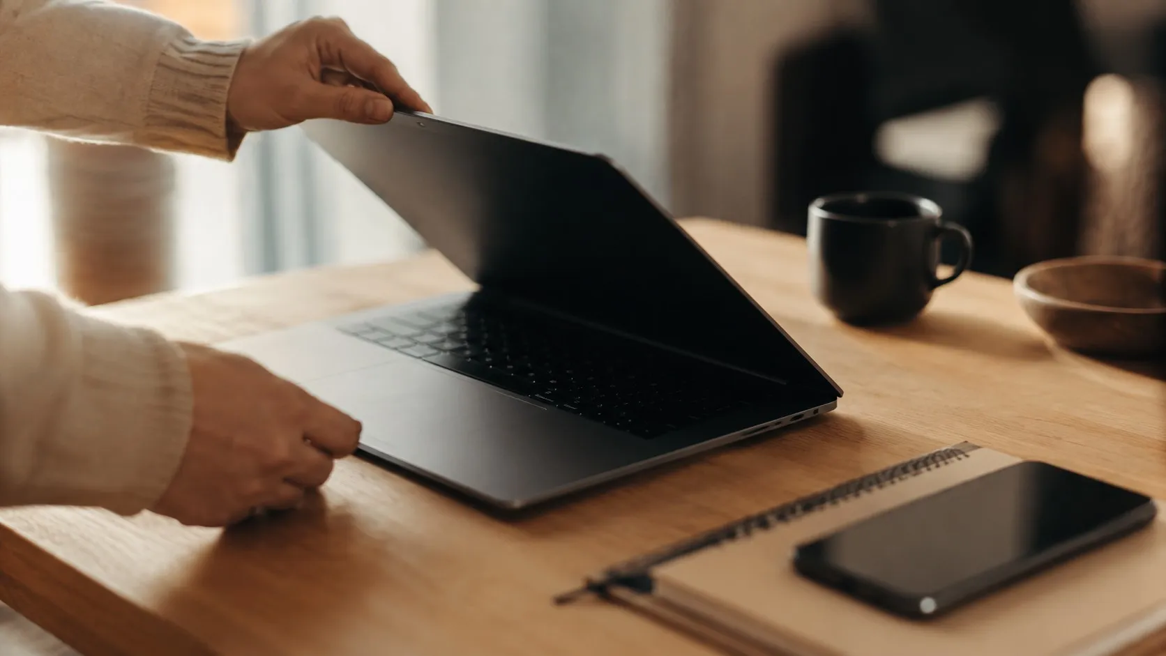 Ett par händer fäller upp en laptop på ett skrivbord med en mobiltelefon bredvid
